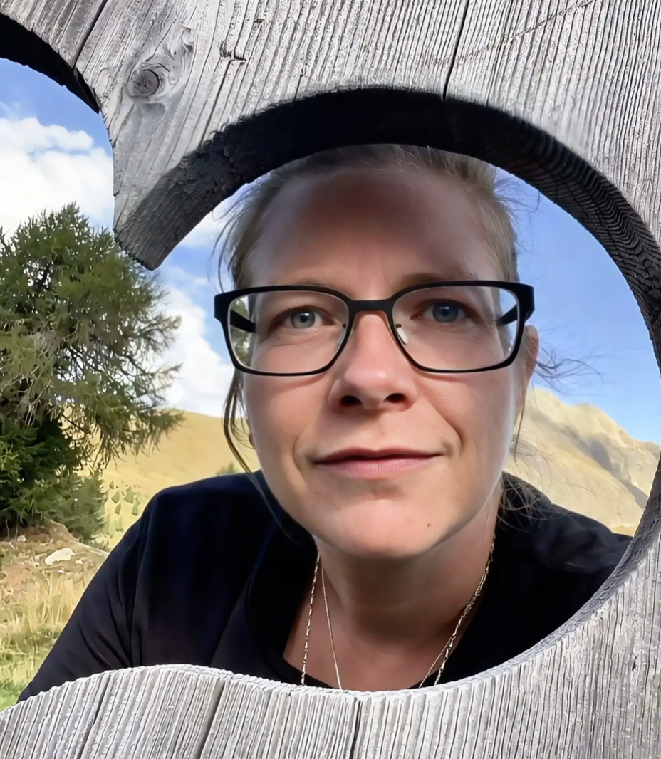 Porträt einer Frau mit Brille unter einem großen Holzherz, im Hintergrund Berge und Himmel.