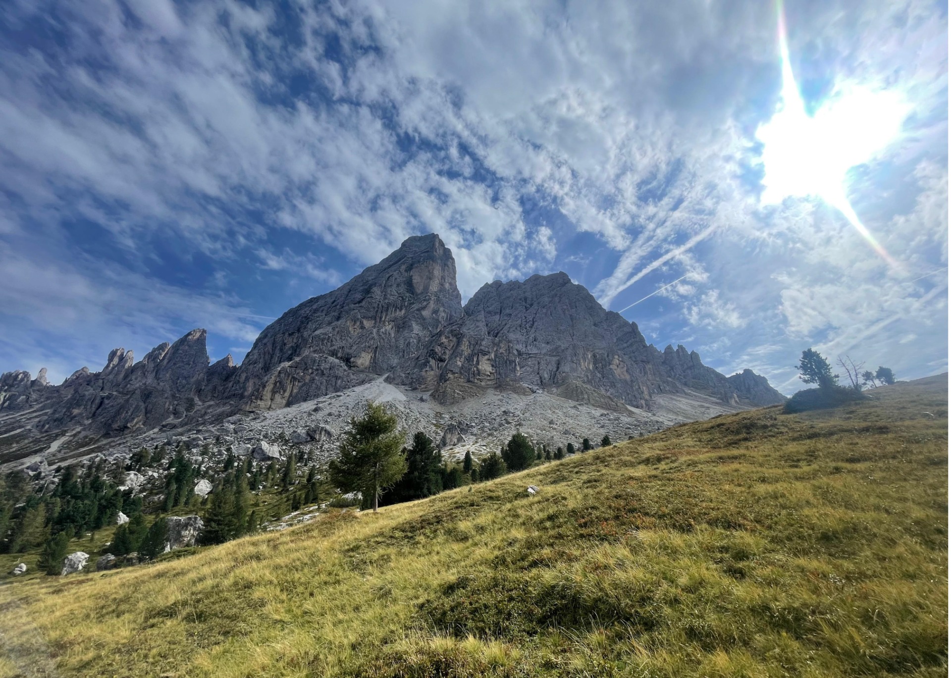 Bergmassiv mit schroffen Felsen, grünen Wiesen im Vordergrund und dramatischem Himmel mit Sonnenstrahlen.