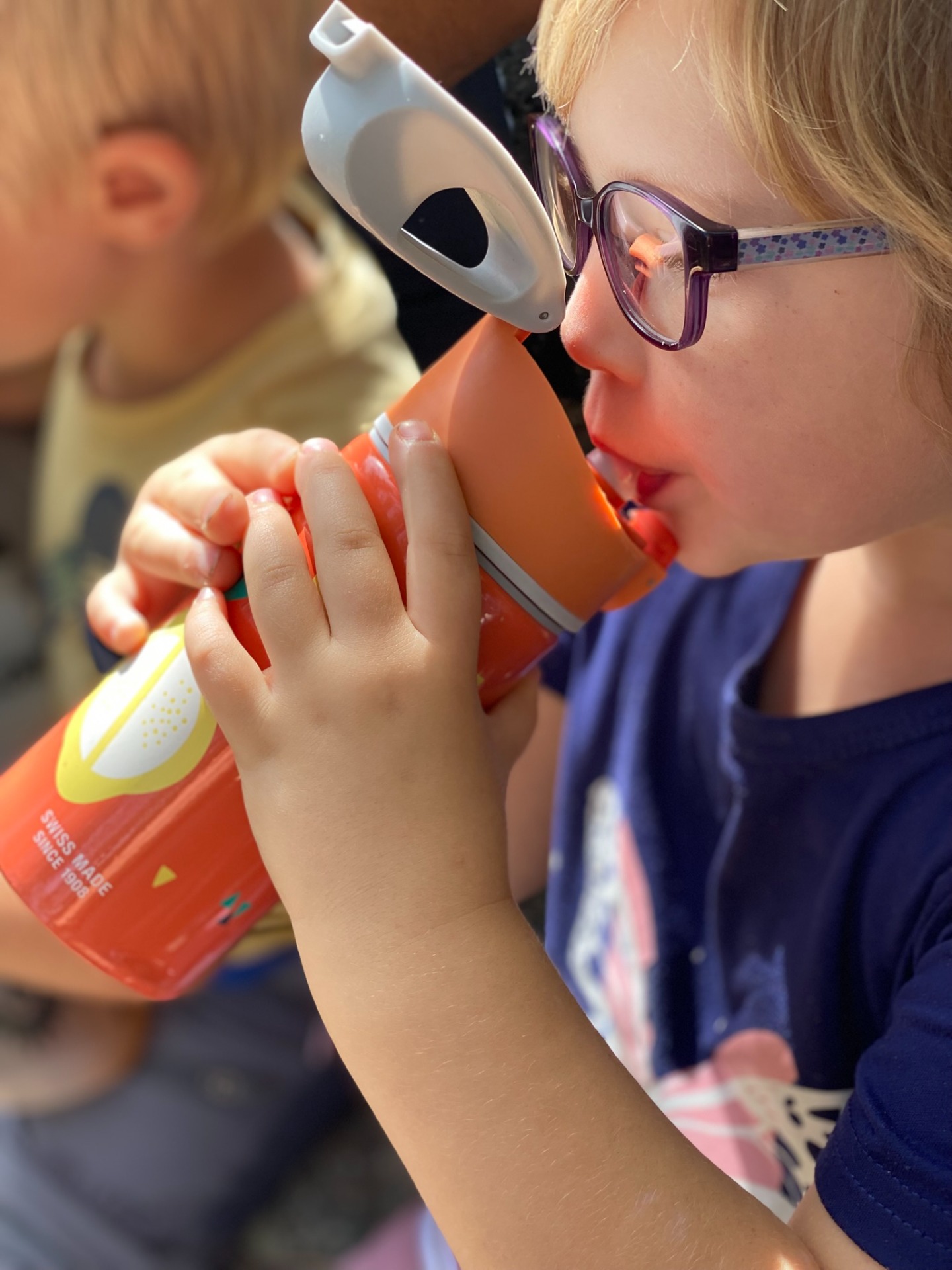 Kind mit Sehbrille trinkt aus einer orangen Trinkflasche von SIGG während ein weiteres Kind im Hintergrund sitzt.