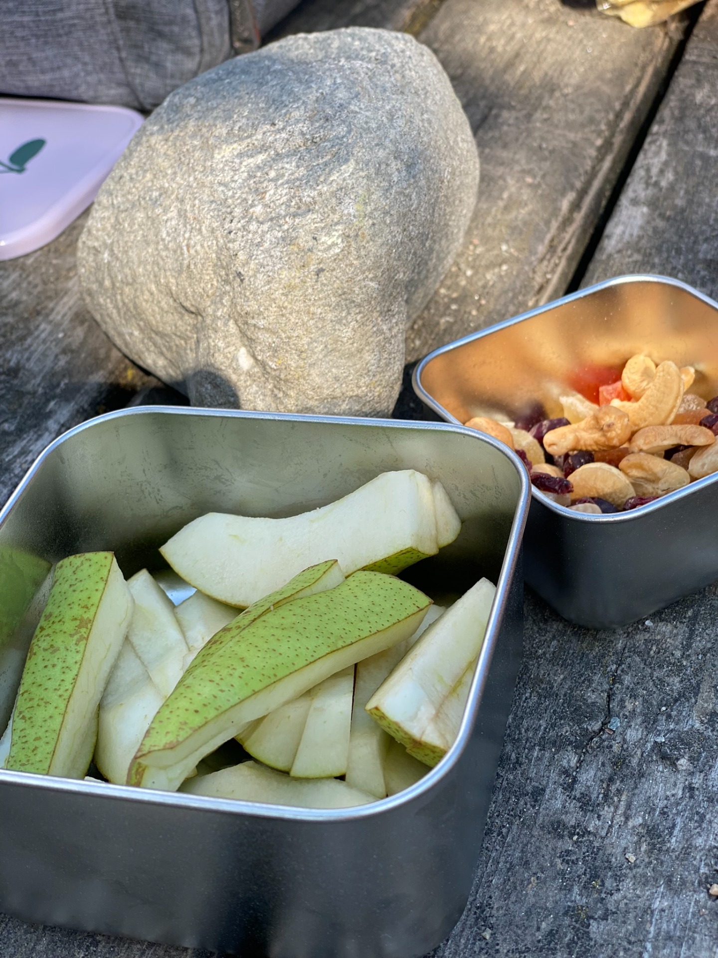 Zwei Metallboxen mit geschnittenen Birnen und Obst, daneben Nüsse und Trockenfrüchte auf grauem Tisch. Snackbox oder Brotdosen von Lässig.