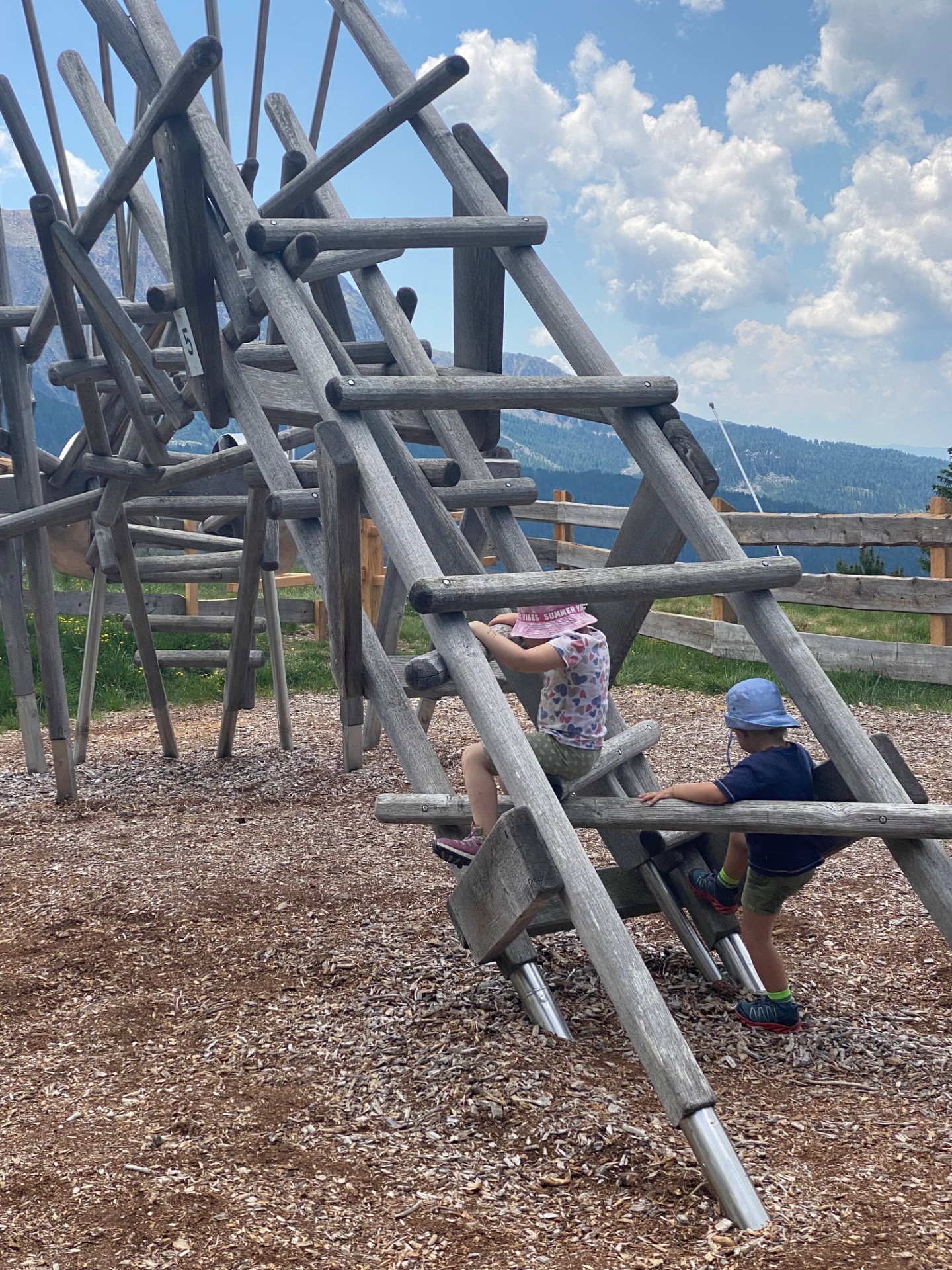 Holzspielgerät mit schrägen Balken, zwei Kinder sitzen darunter, im Hintergrund Berge und blauer Himmel mit Wolken.